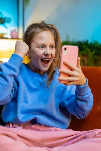 Happy child girl use mobile smartphone typing playing browsing found out great big surprise, good positive news, celebrate online game win. Teenager kid at evening home in room sits on sofa. Vertical