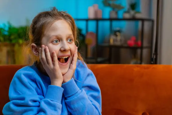 Oh my God, Wow. Caucasian surprised preteen school girl looking at camera with big eyes, shocked by sudden victory good win news celebrating. Excited amazed child kid at home room on couch. Copy-space