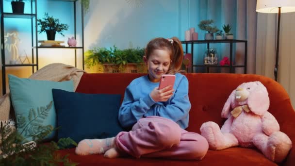 スマートフォンのソーシャルメディアアプリケーションでメッセージのコンテンツをオンラインで共有し リラックスした映画を見ています 女性のティーンエイジャーの子供は 夜の部屋で家で携帯電話を使用してソファに座っています — ストック動画