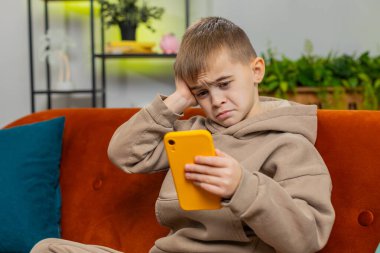 Çocuk akıllı telefon kullanıyor, oyun oynuyor, beklenmedik piyango sonuçlarını kaybediyor, kötü haber, servet kaybı, başarısız eğitim sınavı. Oturma odasındaki küçük erkek çocuk kanepede oturuyor.