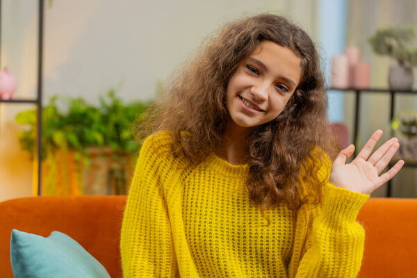 Happy child girl smiling friendly at camera and waving hands gesturing hello, hi, greeting or goodbye, welcoming with hospitable expression. Teenager Caucasian kid at home in living room sits on couch