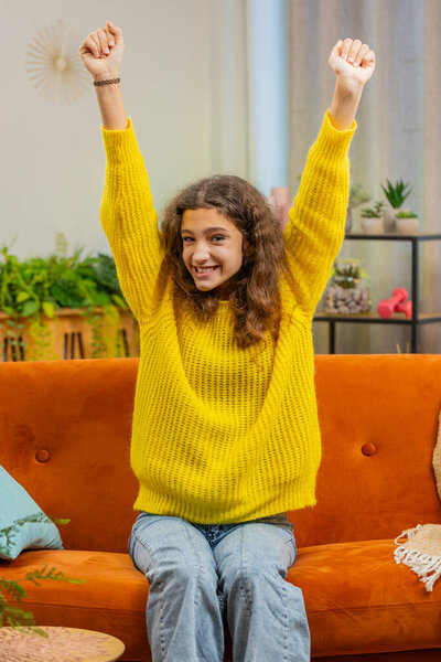 Happy child girl celebrating success, winning, goal achievement good news, lottery luck victory, watching TV championship at home apartment. Teen 14-15 years kid in living room sits on couch. Vertical