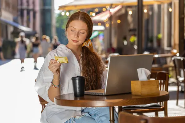 Girl using credit bank card laptop computer while transferring money, purchases online shopping, order food delivery booking hotel room outdoors. Young woman tourist sitting at table in city cafeteria