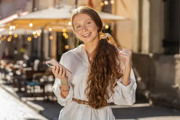 Teenager redhead adult girl use mobile smartphone celebrating win good message news, lottery jackpot victory, giveaway online outdoors. Happy woman tourist walking in urban city street. Lifestyles