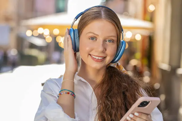 Happy relaxed overjoyed teenager young adult girl in wireless headphones choosing, listening favorite energetic disco rock n roll music dancing outdoor. Child looking at camera on city sunshine street