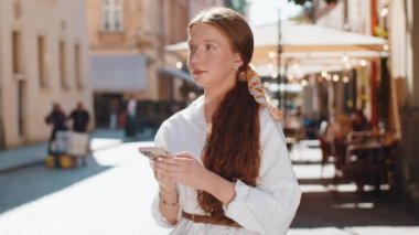 Mutlu gülümseyen kızıl saçlı genç kız internet sosyal medya uygulamalarına girip dışarıda sohbet ederek akıllı telefon yazıları yazıyor. Şehir caddesinde yürüyen çocuk turistler. Yaşam Tarzları