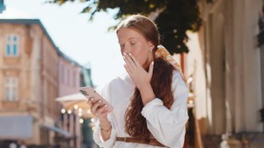 Genç kızıl saçlı kız cep telefonuyla kutlama yapıyor. İyi haber kazan, piyango zaferi kazan, açık havada internetten bağış yap. Şehir caddesinde yürüyen mutlu genç kadın turist. Yaşam Tarzları