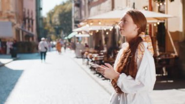 Gezgin, kızıl saçlı, akıllı telefon kullanarak dışarıda gezici navigatör uygulamasında haritada arama yapmak için mesaj yazan bir kız. Şehir merkezinin arka planından geçen çocuk yürüyüşü