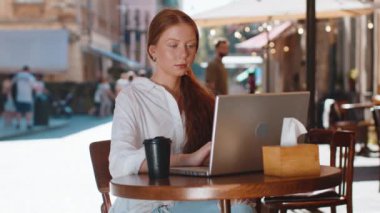 Kızıl saçlı kız eğitim alanında çalışıyor. Sunny Street Cafe 'de uzak bir yerde bilgisayarla çalışıyor. Tatil sırasında açık havada internet sitesinde sohbet ediyor. Gülümseyen kadın turist kameraya bakıyor.