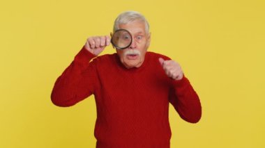 Araştırmacı bilimadamı kıdemli yaşlı adam elinde büyüteçle kameraya bakıyor, aranıyor, analiz ediyor. Sarı arka planda yaşlı büyükbaba