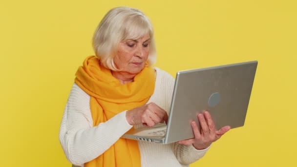 ラップトップコンピュータにタイピングし プロジェクトに取り組み 結果を楽しんで 宝くじのゲームを勝ち取り 成功したオンラインショッピング 勝利を祝う幸せなシニア女性 黄色の背景にある高齢のおばあちゃん — ストック動画