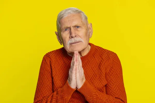 Please, God help. Senior old man praying, looking at camera and making wish, asking with hopeful imploring expression, begging apology, meditation. Mature grandfather isolated on yellow background