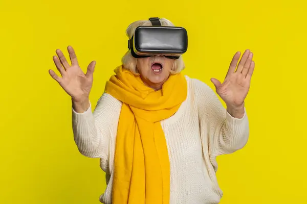 Excited happy senior woman using headset helmet app to play simulation game. Watching virtual reality 3D 360 video. Mature grandmother in VR goggles isolated on yellow background. Future technology