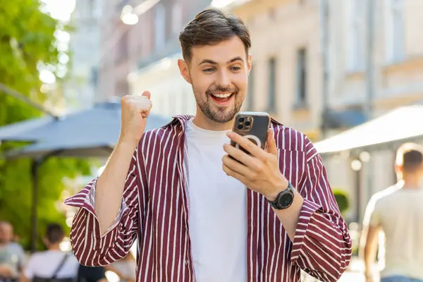 Excited bearded man use mobile smartphone celebrating win good message news, success, lottery jackpot victory, giveaway online outdoor. Happy young guy tourist walking on urban city street. Lifestyles