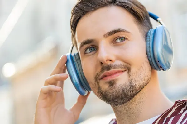 Portrait face of happy relaxed Caucasian bearded young adult man in wireless headphones choosing listening favorite energetic disco rock n roll music dancing outdoor. Guy standing on urban city street