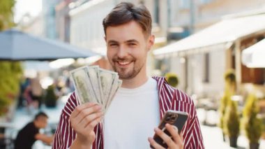 Sakallı mutlu adam elinde bir dolar nakit para tutuyor, akıllı telefon hesap makinesi uygulaması kullanıyor, hediye sipariş etmeyi planlıyor ve internetten otel odası ayırtmayı planlıyor. Esmer genç adam. Şehir sokaklarında turist.
