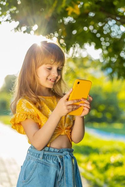 Preteen child kid using smartphone typing text answering messages chatting online looking mobile screen social media app, playing game. Pretty blonde girl standing on city sunny park street. Vertical