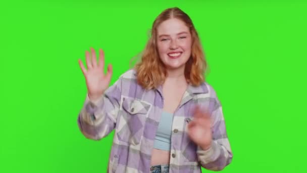 カメラでフレンドリーに微笑む若い白人女性 招待状ホスピタリティな表現で歓迎する挨拶やさよならを振る手 緑色のクロマのキーバックで隔離された赤毛の少女 — ストック動画