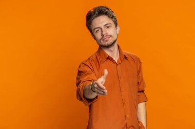 Dost canlısı, beyaz bir adam kameraya uzanmış el sıkışma teklif ediyor. Nazik bir gülümsemeyle selamlıyor. Dayanışma ve güven göstergesi olarak karşılıyor. Genç adam turuncu arka planda kapalı alanda izole edilmiş.