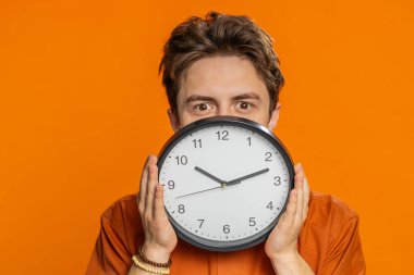 Ofis saatini arkasına saklayan çok katı bir beyaz adam, vardiyayı kontrol ediyor, işe geç kalıyor, son teslim tarihini geciktiriyor, yüzünü gizliyor, casus. Turuncu arka planda izole edilmiş bir adam.