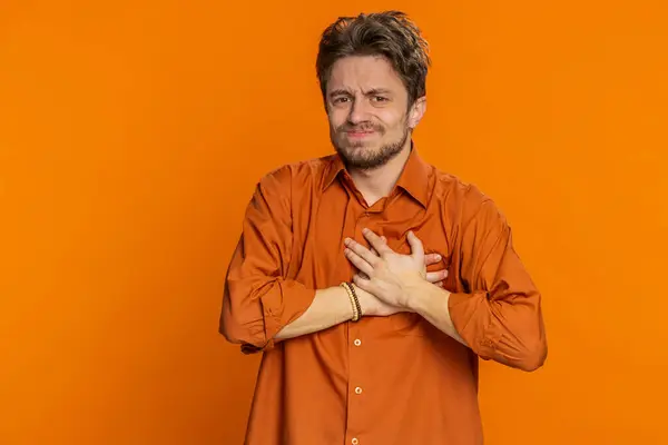 Stressed scared sick ill Caucasian man suffers from tension holding chest with heart pain attack risk of stroke pressure symptom, feeling bad sick ill. Young guy isolated on orange background indoors