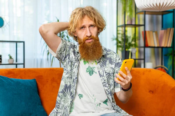 Sad displeased man use smartphone typing browsing, loses becoming surprised sudden lottery game results, bad news, fortune loss, fail, deadline, virus. Young Caucasian guy at home living room on couch