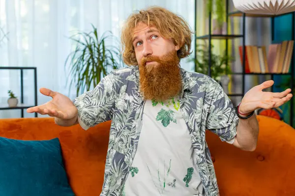 I dont know what to say. Confused Caucasian redhead man feeling embarrassed about ambiguous question, having doubts, no idea, being clueless and uncertain. Young bearded guy at home room on couch