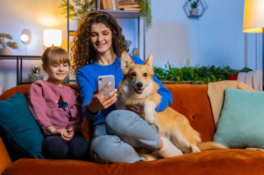 Kanepede akıllı telefon kullanarak Corgi köpeğiyle selfie çeken neşeli anne ve kız çocuğu. Mutlu aile paylaşımı sosyal medya çevrimiçi video görüşmesi hafta sonları evdeki oturma odasında koltukta oturur.
