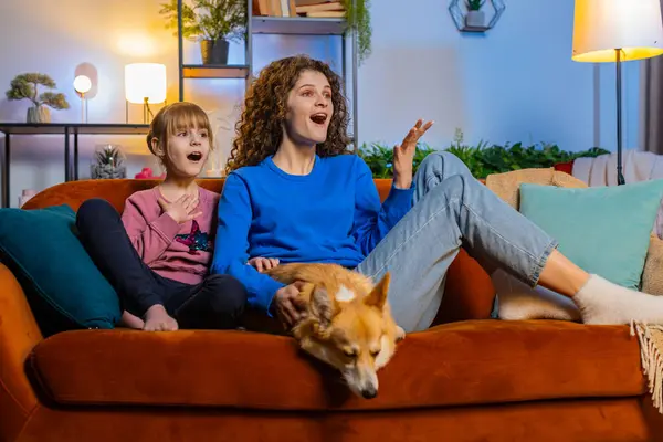 Surprised mother and daughter child girl sitting beside corgi dog watching TV in night room at home. Happy family in casual clothes watching favorite TV show sits on sofa in apartment during weekend.