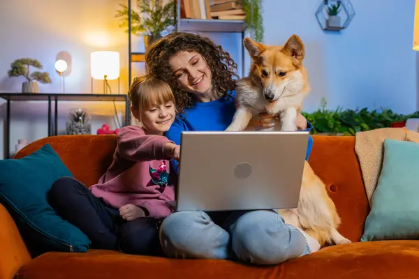 Smiling mother and daughter child girl using laptop sitting beside cute corgi dog on couch in living room. Family watching movie together on netbook during weekend at home. Online education concept.