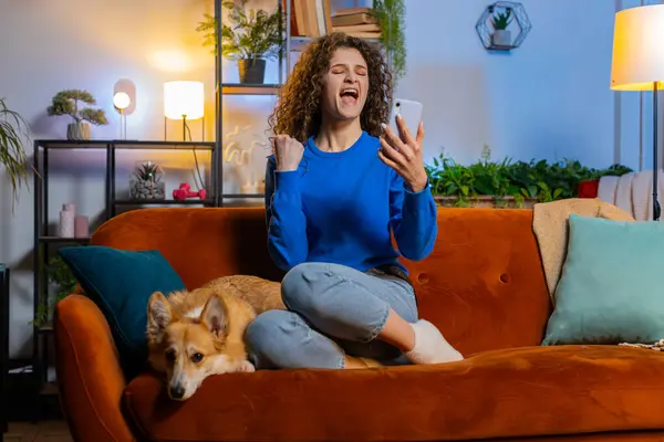 Surprised young woman with curly hair browsing smartphone winning lottery sitting beside corgi dog on sofa in living room at home. Excited female girl celebrating success clenching fists on sofa.