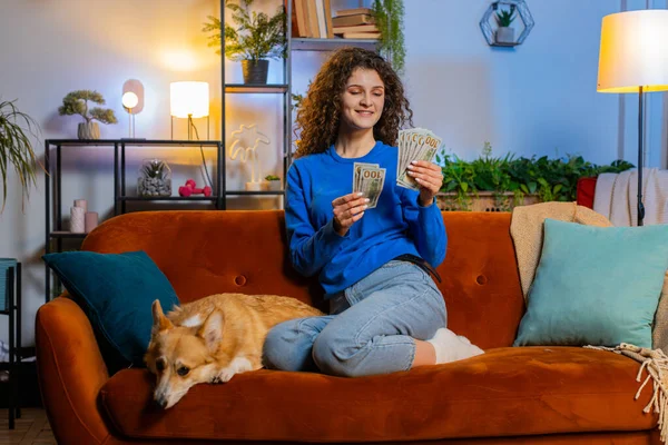 Successful happy rich woman counting dollar banknotes while sitting on sofa beside corgi dog in living room at home. Smiling girl calculating budget, savings, earnings, planning vacation in apartment.