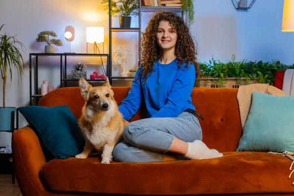Happy young woman with curly hair in casual clothes caressing cute corgi dog relaxing on comfortable sofa in living room at home. Beautiful girl lady spending leisure time with pet dog in apartment