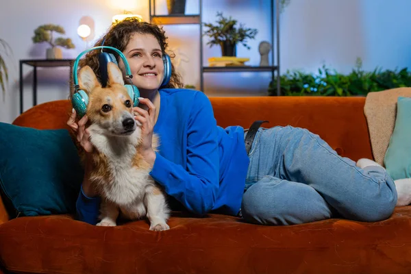 Happy young woman listening to music through wireless headphones lying on couch with corgi dog. Cheerful female girl with curly hair dancing on music in living room at home. Enjoying dance music