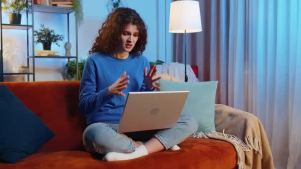 怒气冲冲的年轻女性卷发使用笔记本电脑上网本接收坏消息 而坐在客厅的沙发上 不高兴的女自由撰稿人在公寓的沙发上坐立不安 — 图库视频影像