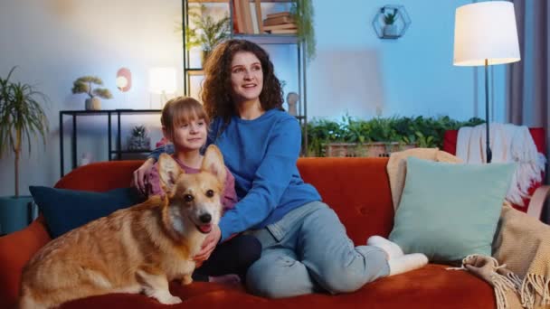 令人惊讶的是 母亲和女儿的女儿坐在科吉狗旁边 在客厅里看电视 穿着休闲装的快乐家庭坐在沙发上看最喜欢的电视节目 — 图库视频影像