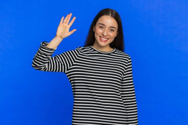 Kameraya gülümseyen beyaz bir kadın, el sallayarak merhaba diyor ya da davetkar bir yüz ifadesiyle veda ediyor. Güzel kız mavi stüdyo arka planında izole edilmiş.