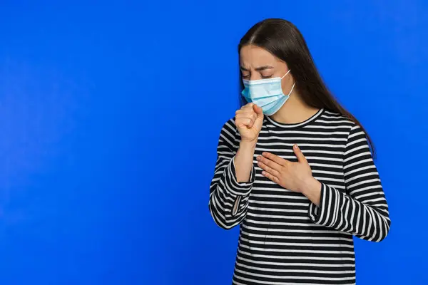 Unhealthy young woman coughing covering mouth with hand, feeling sick, allergy or viral infection symptoms, fever pain virus. Pretty brunette girl isolated alone on blue studio background. Copy-space