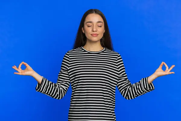 Keep calm down, relax, inner balance. Young woman breathes deeply with mudra gesture, eyes closed, meditating with concentrated thoughts, peaceful mind. Attractive girl isolated on blue background