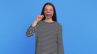 Çizgili bluzlu mutlu beyaz kadın elinde büyüteçle kameraya bakıyor, komik gülümseyen aptal bir yüz ifadesi sergiliyor. Sağlık, hijyen, stomatoloji. Mavi arkaplanda izole edilmiş kız