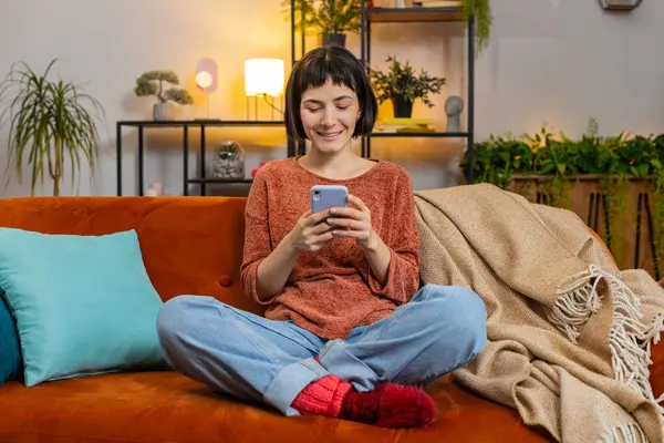 Gündelik giysiler içinde, akıllı telefondan mesaj atan mutlu genç bir kadın. İspanyol kız kanepede oturuyor ve evdeki oturma odasında cep telefonu kullanıyor. Dairede internetten film izleyen bir kadın..