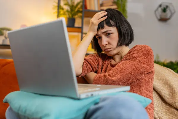 Angry woman using laptop typing browsing working, loses becoming surprised sudden lottery results bad news fortune loss fail computer virus error. Upset Caucasian girl on couch in living room at home.