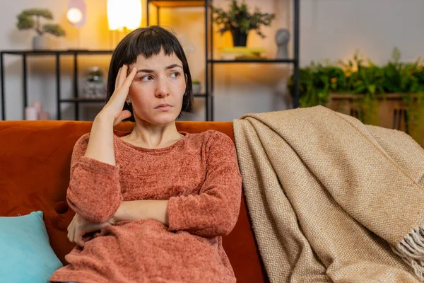 Sad lonely Caucasian girl looks pensive thinks over life concerns suffers from unfair situation sitting on sofa. Upset woman with hand in hair depressed feeling bad annoyed in living room at home