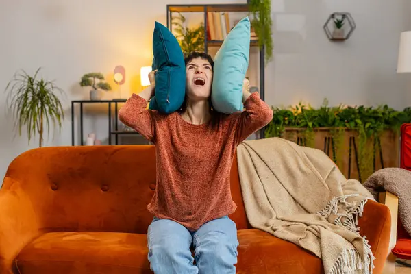 Irritated Caucasian woman girl shouting covers ears with cushions annoyed by noisy neighbors suffers from headache sitting on sofa. Thin walls at home flat without sound insulation repair work concept