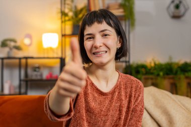 Heyecanlı beyaz kadın portresi onaylayıcı bir şekilde kameraya bakıyor, olumlu, iyi haber, olumlu geri bildirim gibi. Evdeki oturma odasında oturan gülümseyen kız..