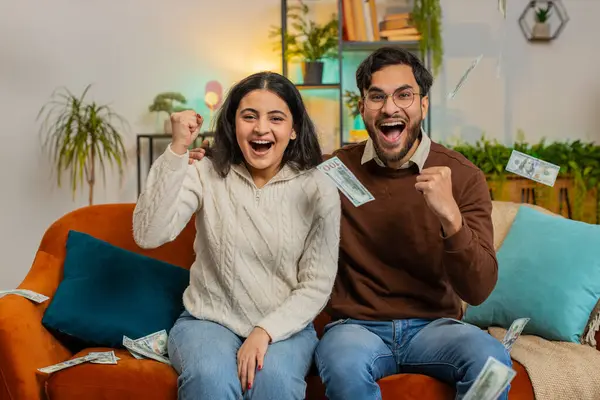 Happy young Indian couple with rising hands fists shouting, celebrating success winning lottery jackpot goal achievement, good news victory. Money cash dollar rain falling on family on sofa at home.