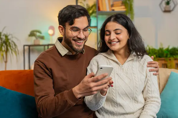 Multiracial excited couple sitting on sofa and swipe smartphone at home. Happy Indian man and woman discussing together spend time, relaxing and enjoying communicate on mobile phone in room apartment