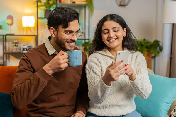 Cheerful young Hispanic couple experiencing fun, amusement as they browse social media applications together on smartphone. Happy girlfriend boyfriend talking on sofa in room drinking tea or coffee