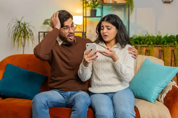 Disappointed multiethnic couple reading together bad news on smartphone while sitting on sofa in room at home. Sad Hispanic family are depressed by unpleasant message shocked by information about loss