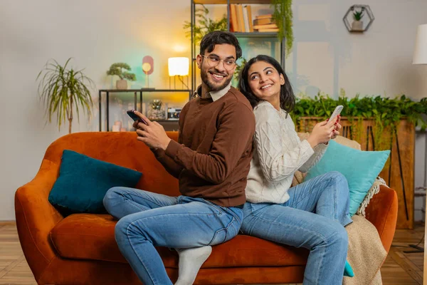 Side view of cheerful diverse couple in casual clothes sitting back to back using smartphones on sofa in living room at home. Young family talking while browsing mobile phone on couch in apartment.